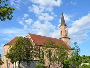 Entdecken Sie Königsbrunn! Historische Kirche mit spitzem Turm und Bäumen unter blauem Himmel