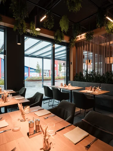 Cervus: Ihre Eventlocation in Augsburg und Umgebung Gemütliches Restaurant mit Holztischen, Stühlen und hängendem Grün neben großen Fenstern
