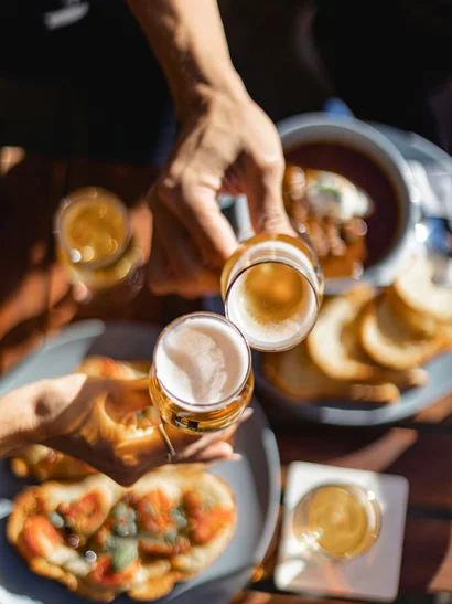 Biergarten rund um Königsbrunn gesucht? Zwei Hände stoßen mit Biergläsern an, Essen auf dem Tisch darunter