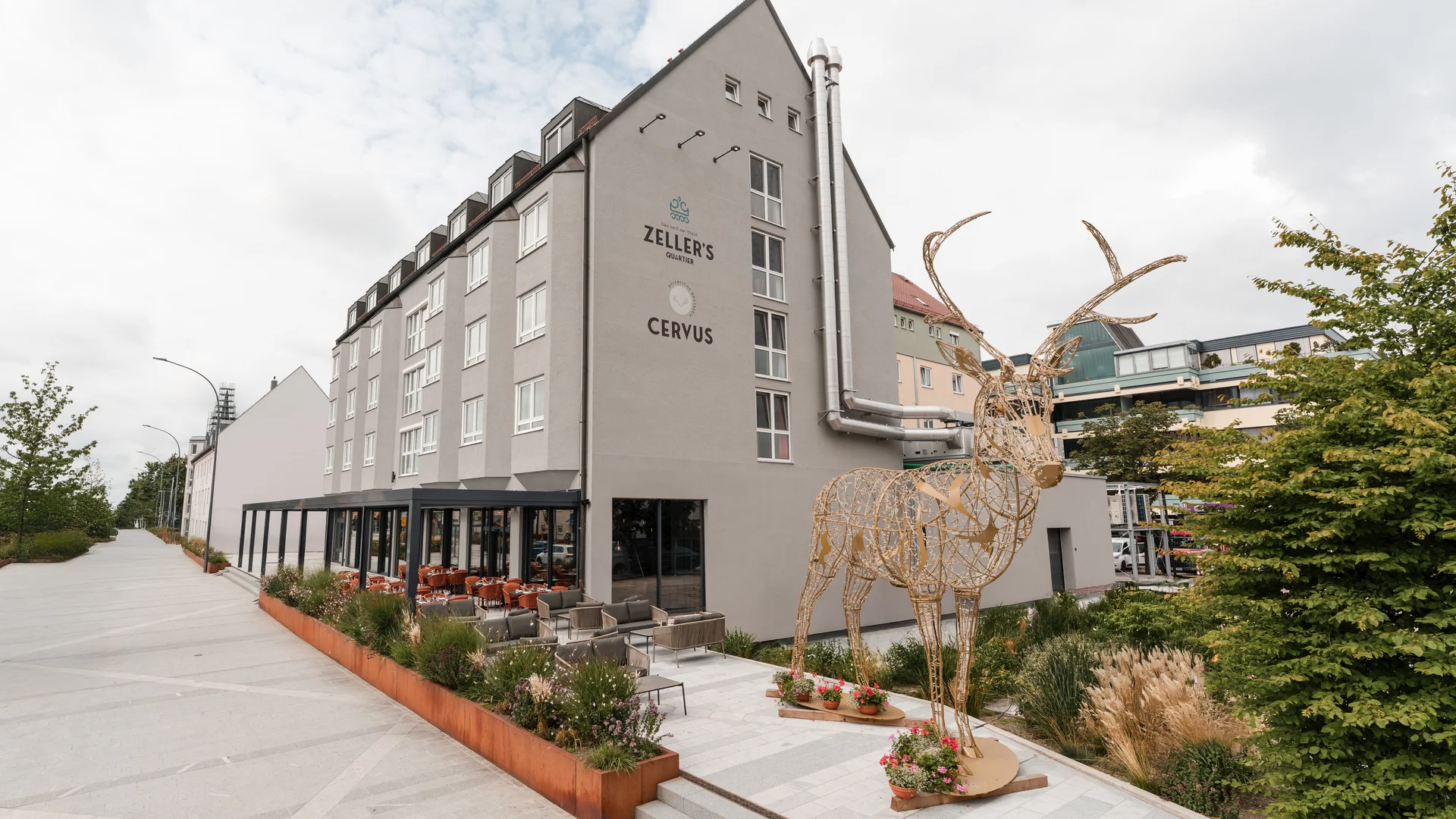 Ihr besonderes Hotel in Königsbrunn: Zeller’s Quartier Modernes Gebäude mit Caféterrasse und großer Hirschskulptur aus Holz im Freien
