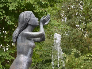 Entdecken Sie Königsbrunn! Bronzeskulptur einer Frau neben einem sprudelnden Wasserbrunnen vor grünem Laub